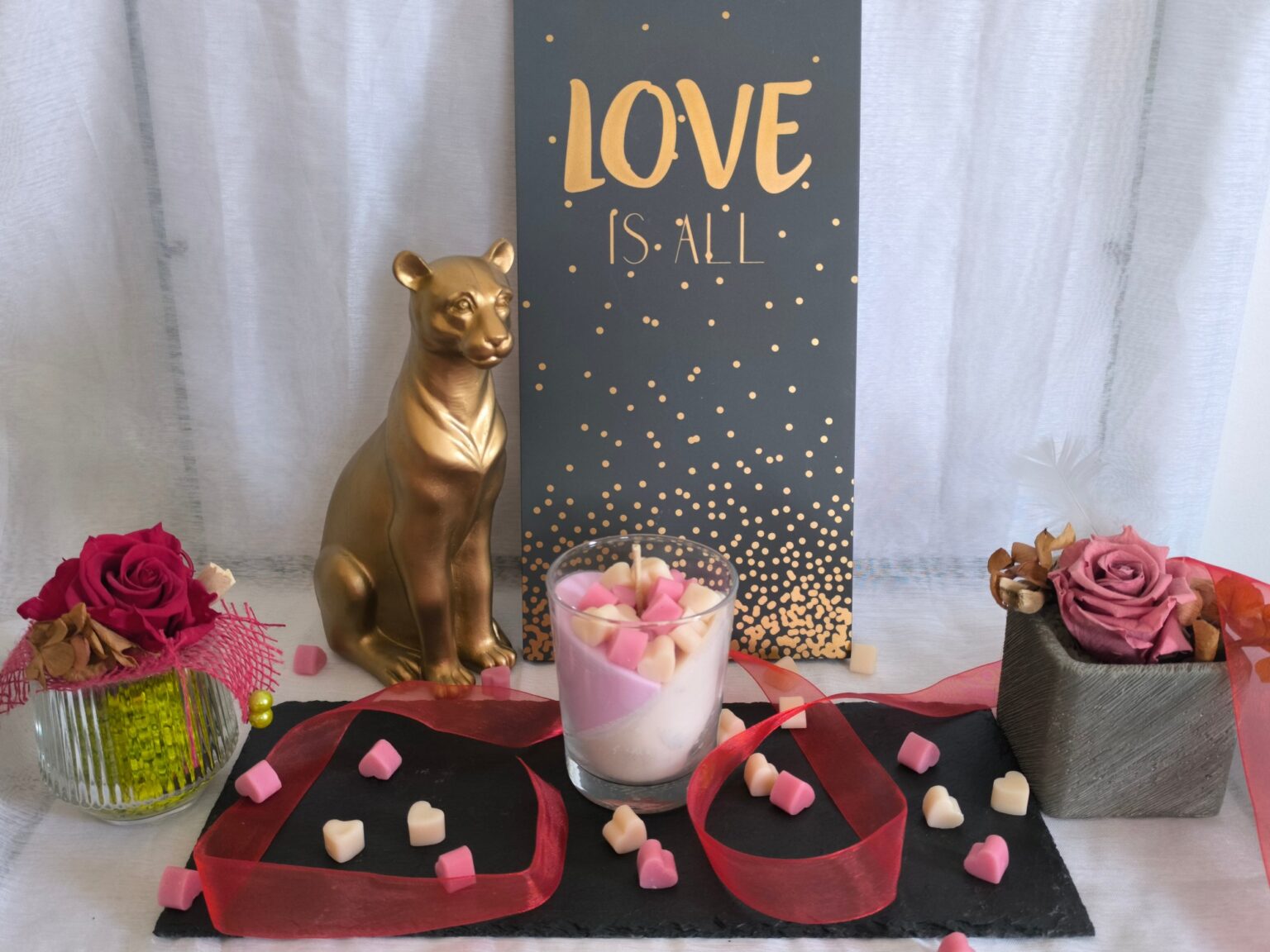 Bougie Be my love dans son verre, garnie de cœurs en cire, sur une ardoise noire entourée de roses éternelles, d'un ruban rouge et d'une panthère dorée