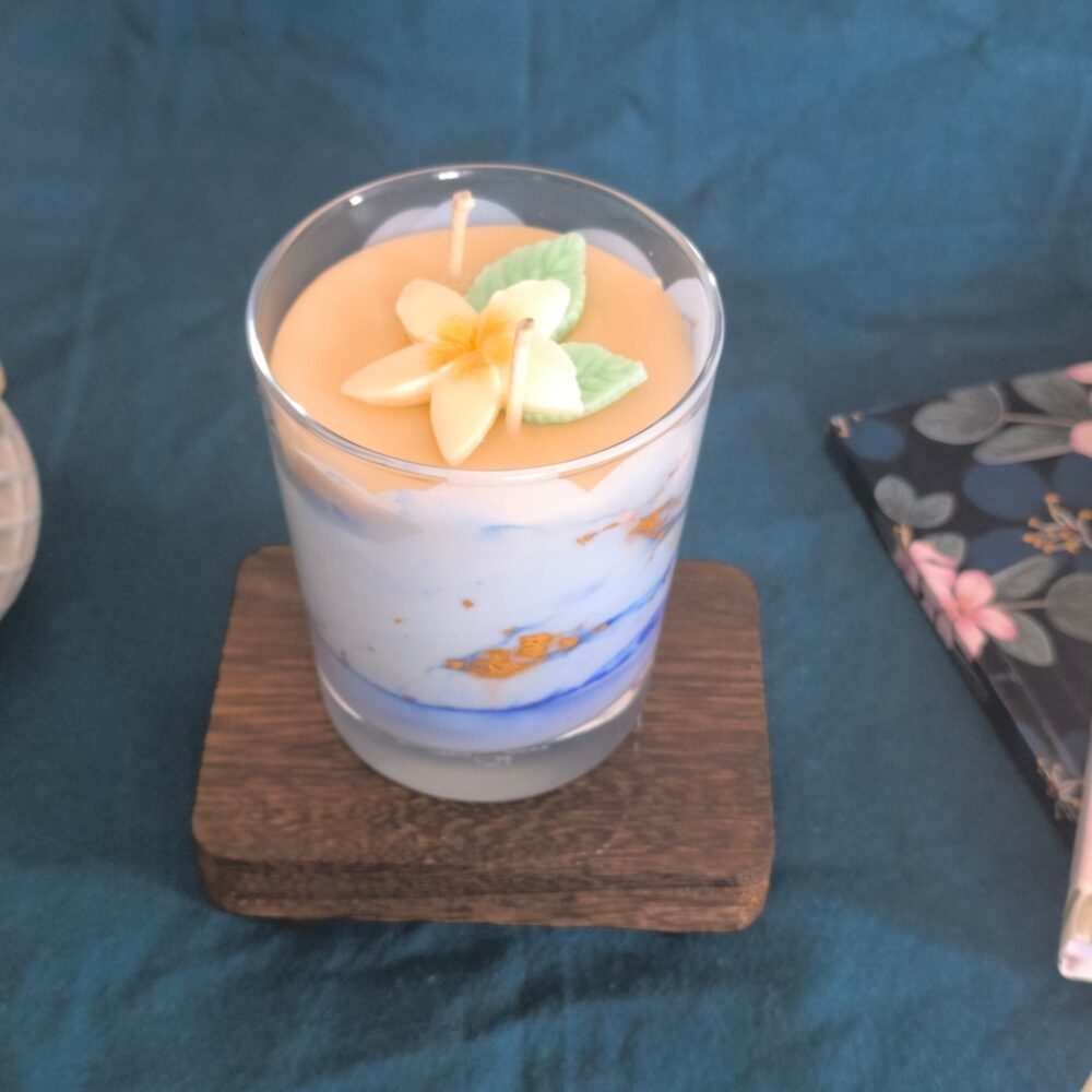 Bougie Rêve Polynésien dans son verre, cire blanche et orange surmontée d'une fleur de frangipanier, posée sur un dessous de verre en bois