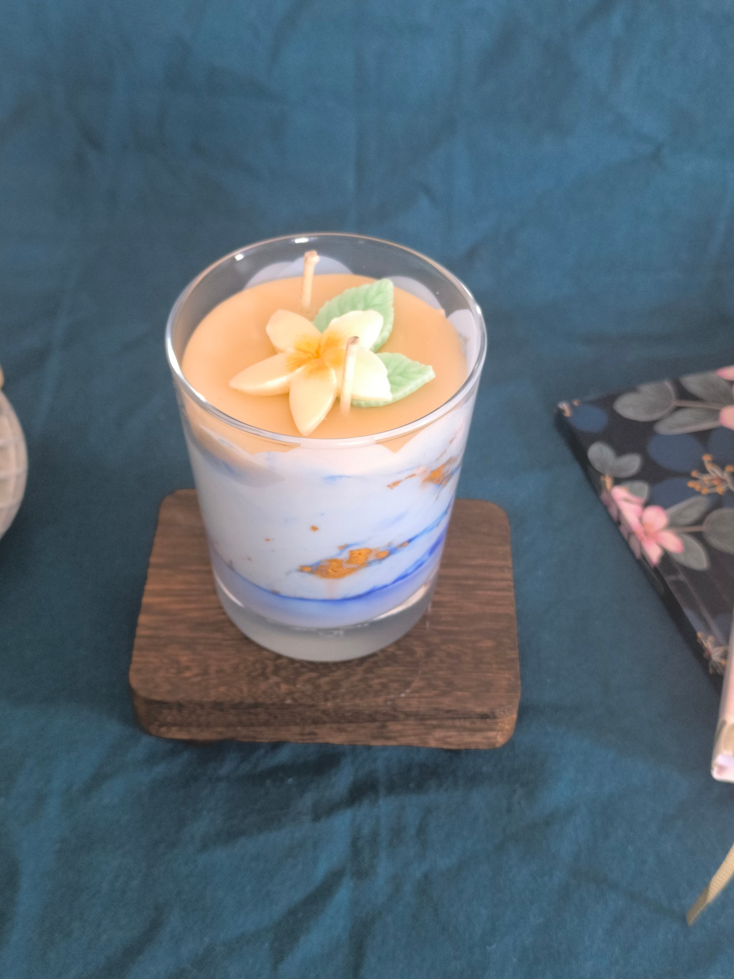 Bougie Rêve Polynésien dans son verre, cire blanche et orange surmontée d'une fleur de frangipanier, posée sur un dessous de verre en bois